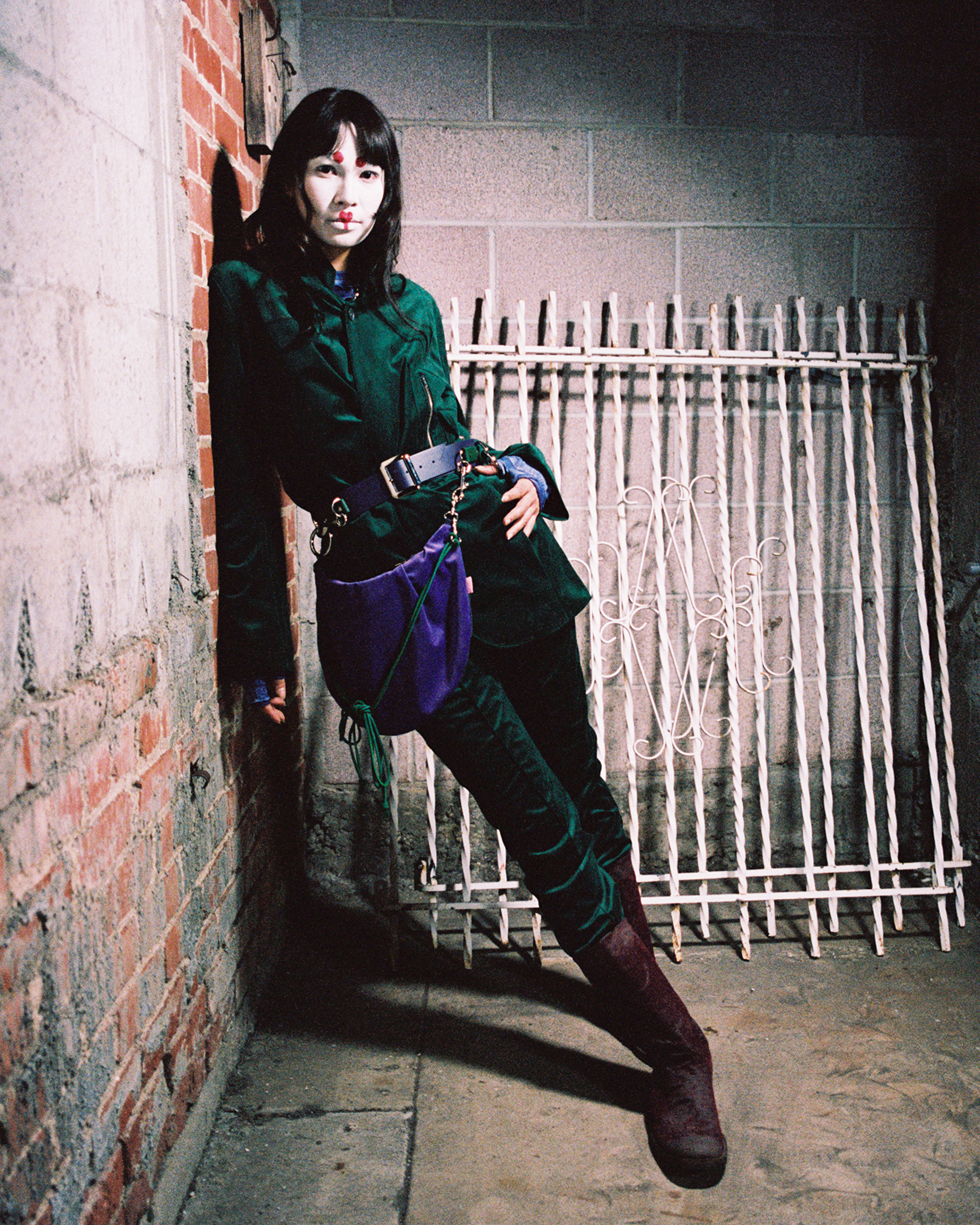 Model with white facepaint with graphic red in a green outfit leaning against a brick wall